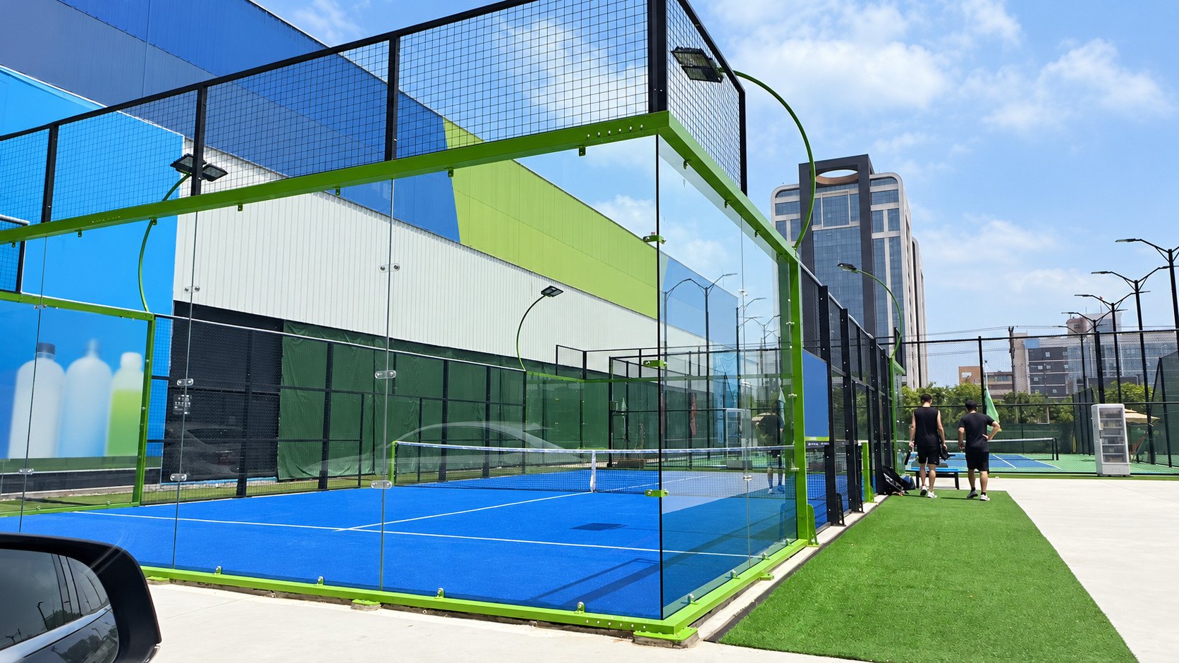 Blue outdoor padel court with a green frame, glass corner and courtside walkway in an urban setting